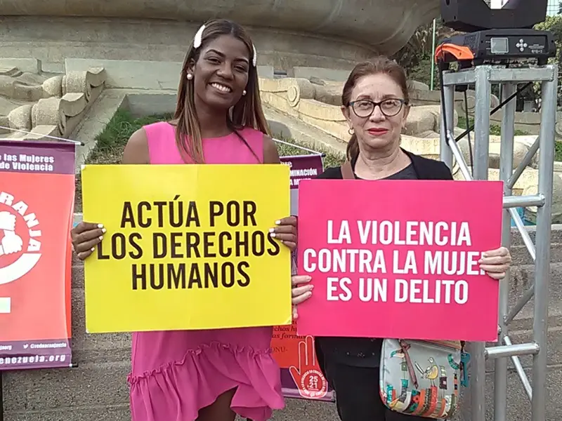 Two women holding signs that read «Actúa por los derechos humanos» (Act for human rights) and «La violencia contra la mujer es un delito» (Violence against women is a crime) at an outdoor event in Caracas